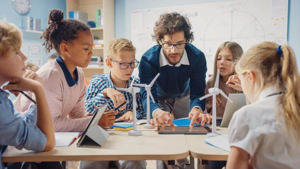 Teacher guiding diverse students in a classroom with wind turbine models and tablets on desks.